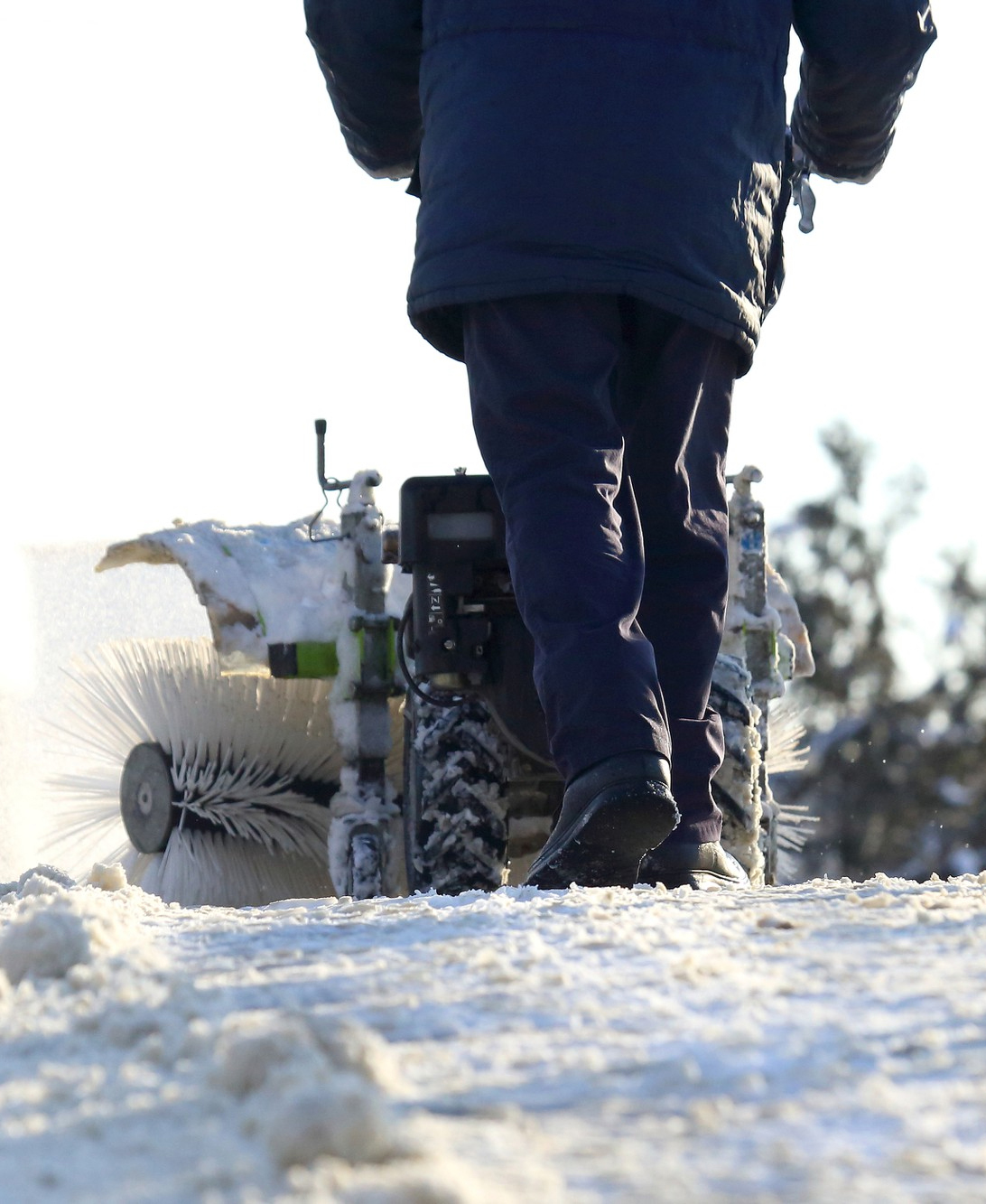 Winterdienst mit Kehrmaschine auf verschneitem Weg Eine Person schiebt eine motorisierte Kehrmaschine über einen schneebedeckten Weg. Die Kehrbürsten drehen sich sichtbar, während Schnee zur Seite geworfen wird. Die Szene spielt bei Tageslicht im Freien.